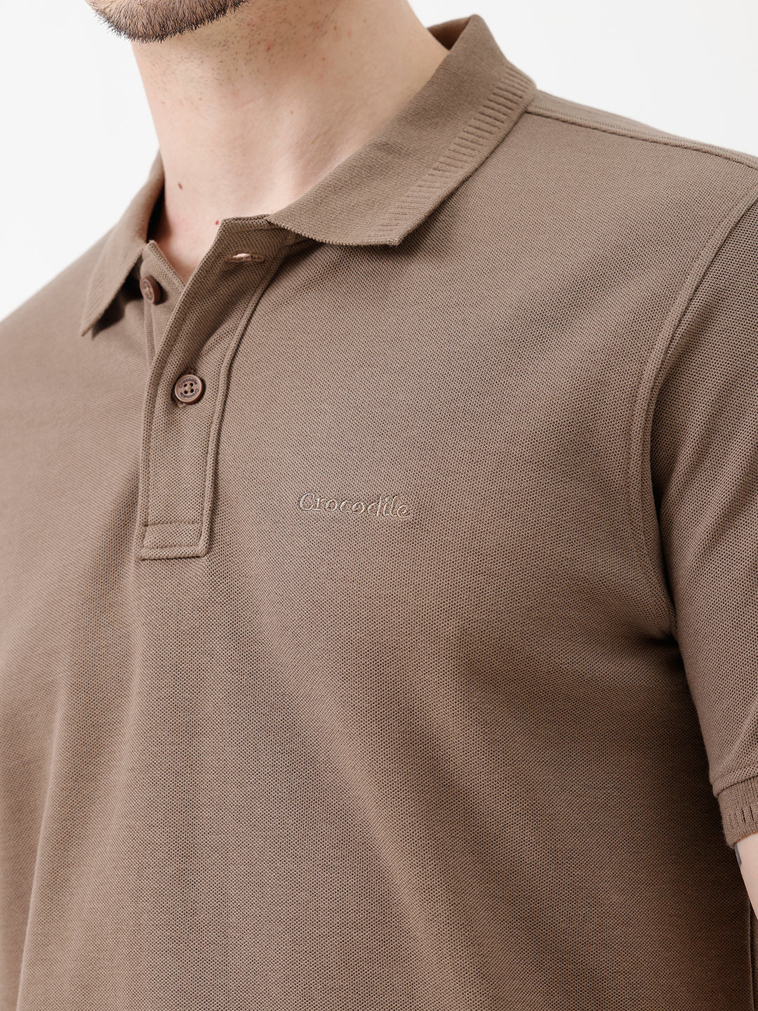 A man wearing a brown short-sleeve polo shirt with dark navy pants, standing against a plain light gray studio background.