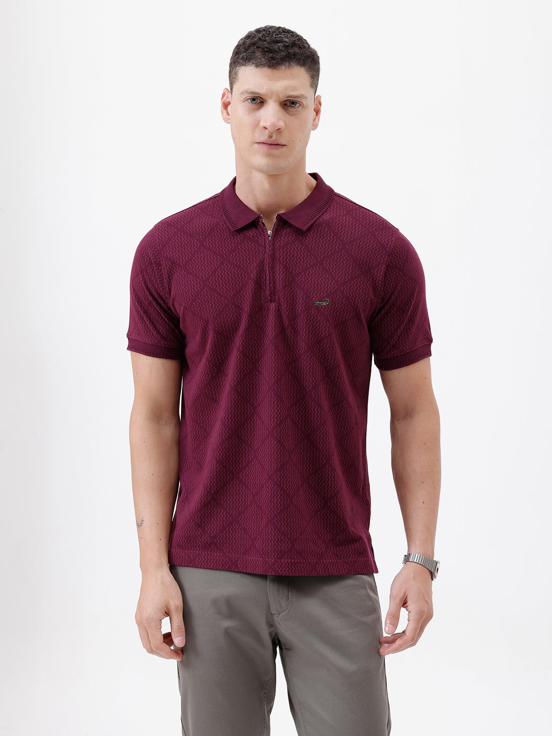A man wearing a short-sleeve polo shirt in deep burgundy with a subtle textured geometric pattern, styled with gray trousers, sunglasses, and a watch against a plain white background.