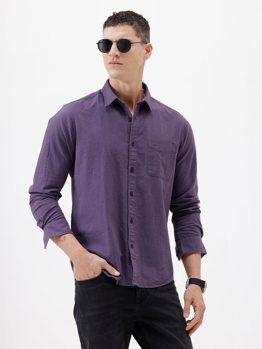 A man wearing a long-sleeve button-down shirt in a muted purple color, paired with black pants, standing against a plain white studio background.