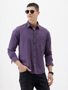 A man wearing a long-sleeve button-down shirt in a muted purple color, paired with black pants, standing against a plain white studio background.
