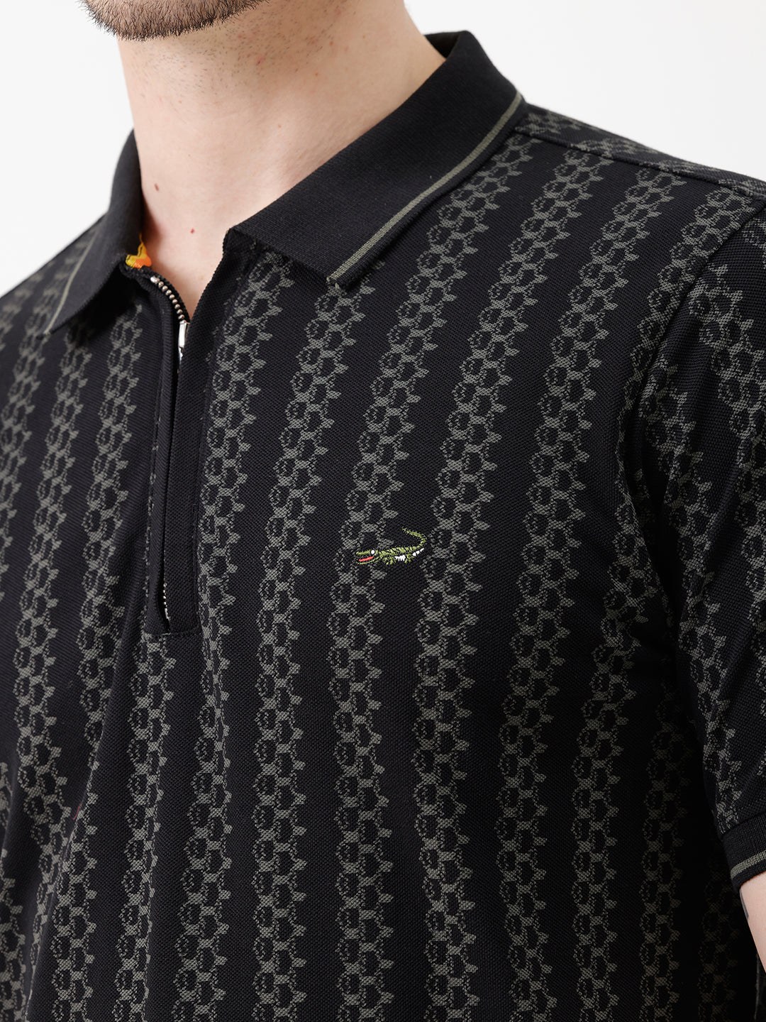 A man wearing a black short-sleeve polo shirt with a subtle geometric pattern, paired with dark gray trousers, standing against a plain white studio background.