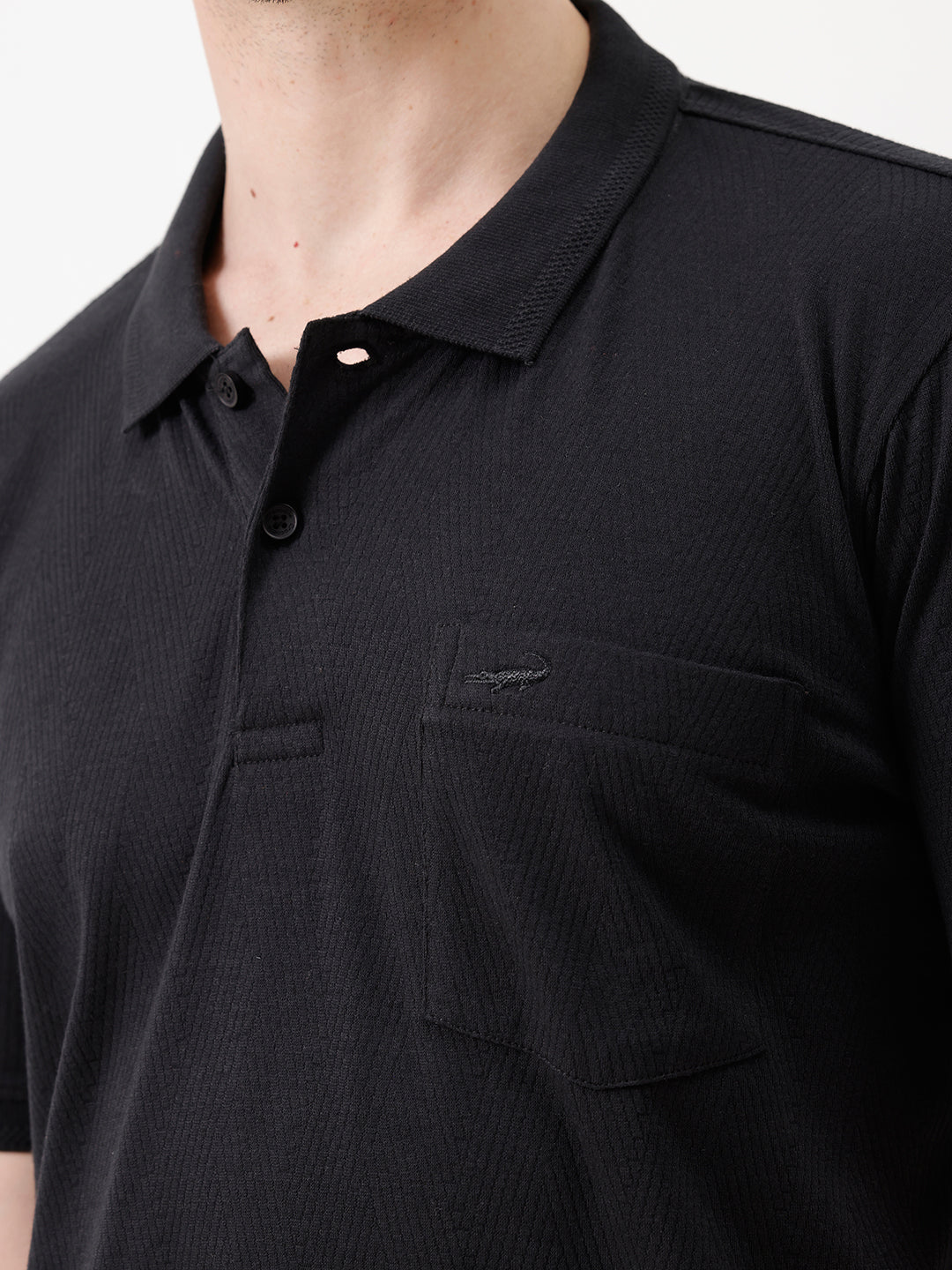 A man wearing a black short-sleeve polo shirt with a single chest pocket, paired with beige trousers, standing against a plain white studio background.
