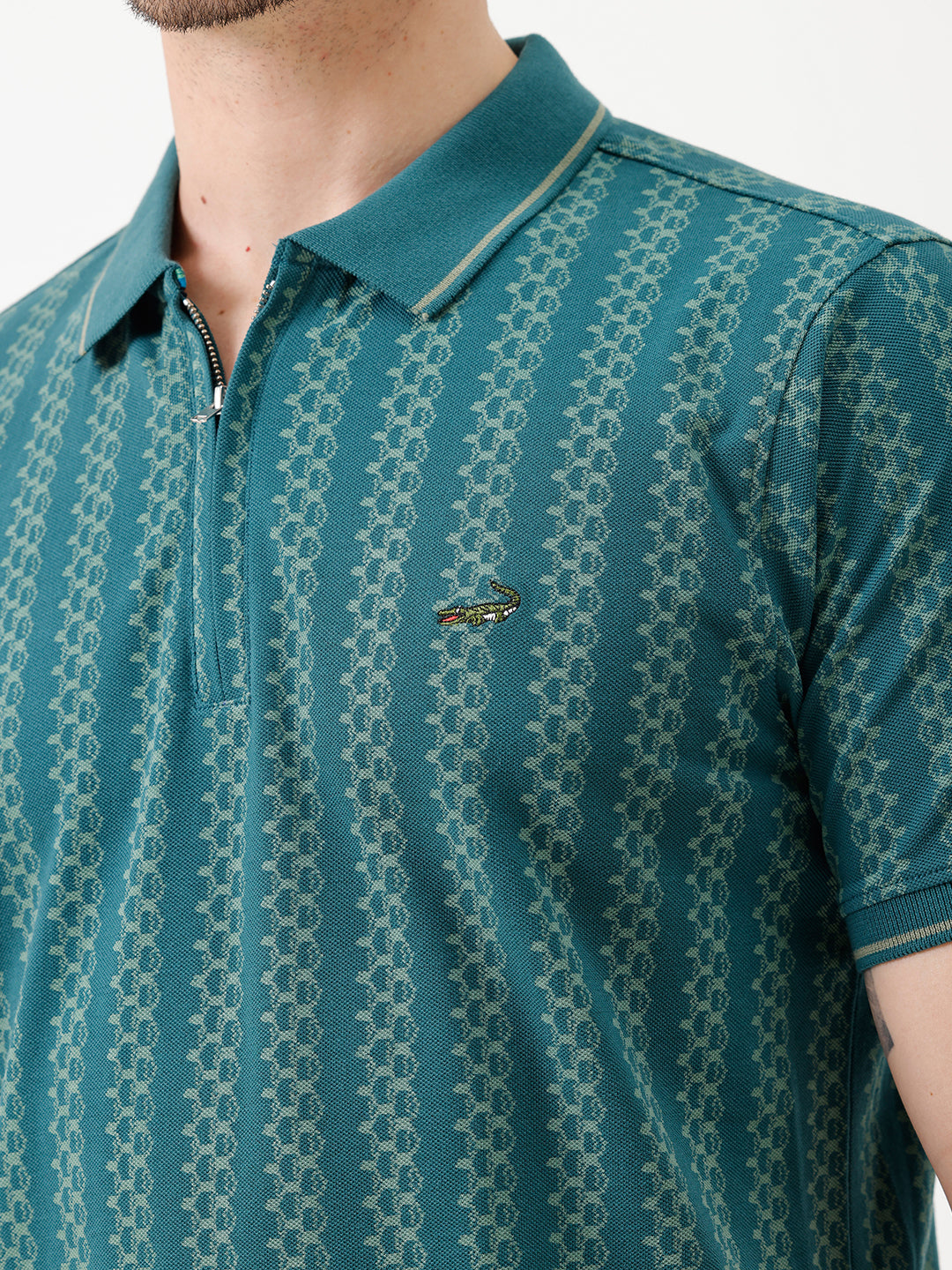 A man wearing a teal green short-sleeve polo shirt with a white geometric pattern, paired with black jeans, standing against a plain white studio background.