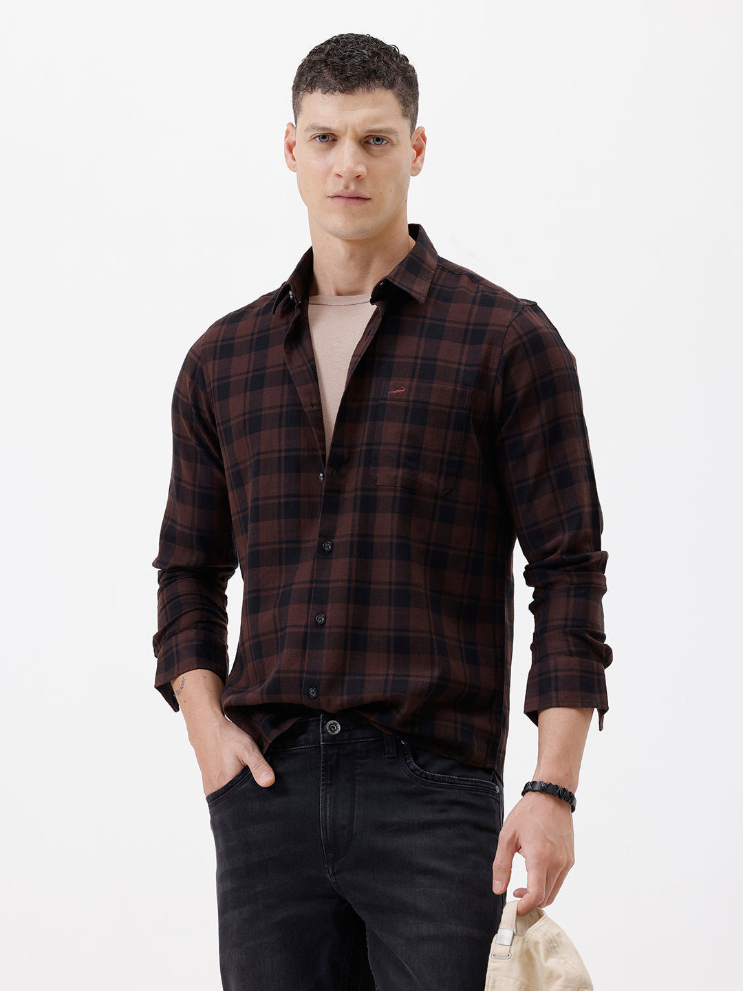 A man wearing a dark brown and black plaid long-sleeve button-up shirt, paired with dark pants, standing against a plain light gray background.