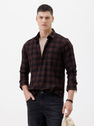A man wearing a dark brown and black plaid long-sleeve button-up shirt, paired with dark pants, standing against a plain light gray background.