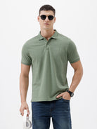 A man wearing a short-sleeve polo shirt in sage green, paired with dark blue jeans, standing against a plain light gray background.