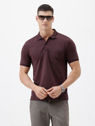 A man wearing a short-sleeve polo shirt in dark burgundy with a chest pocket, paired with gray trousers, standing against a plain light gray background.