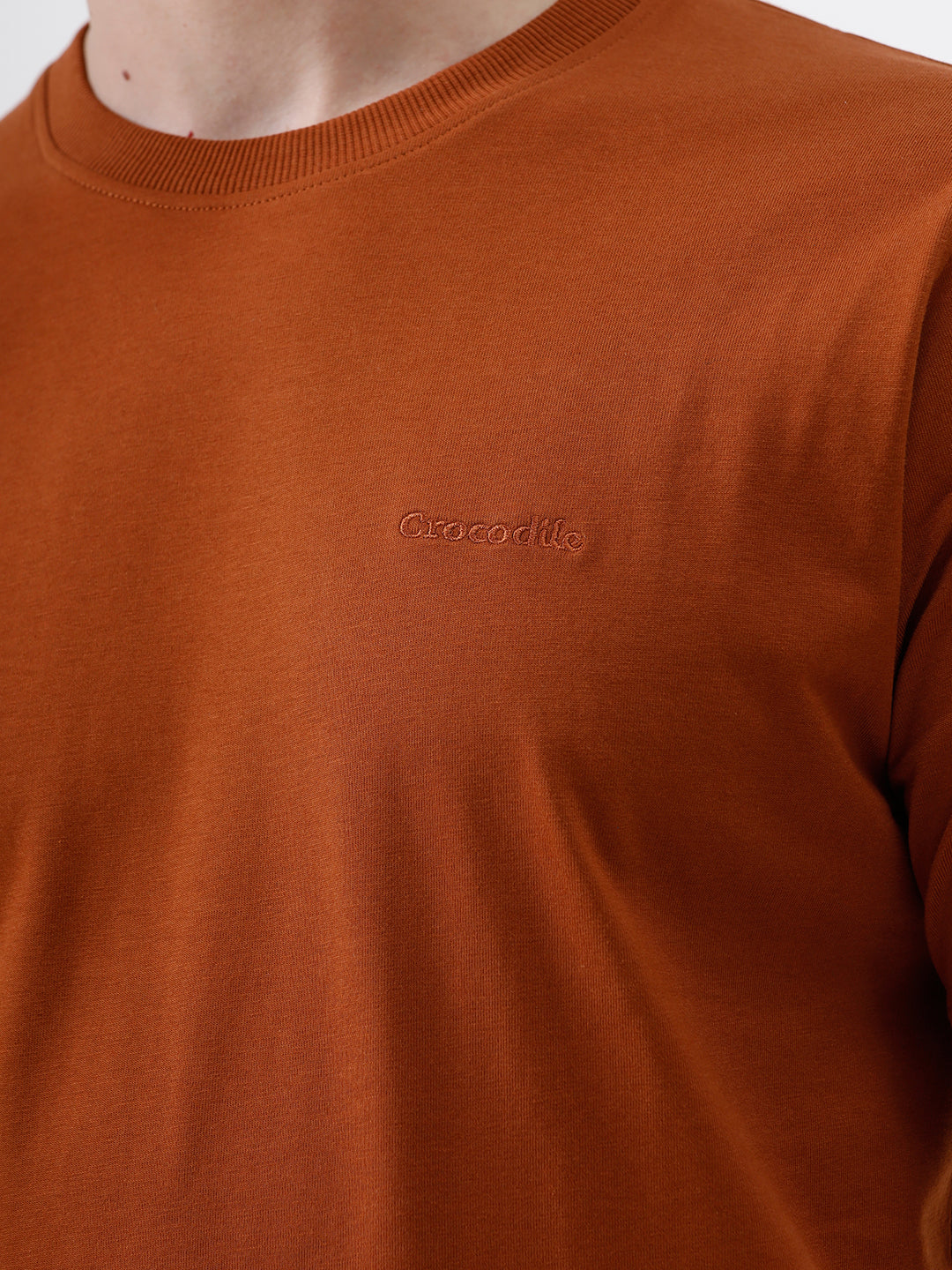 A man wearing a rust-orange crew-neck t-shirt and dark blue jeans, standing against a light gray studio background.