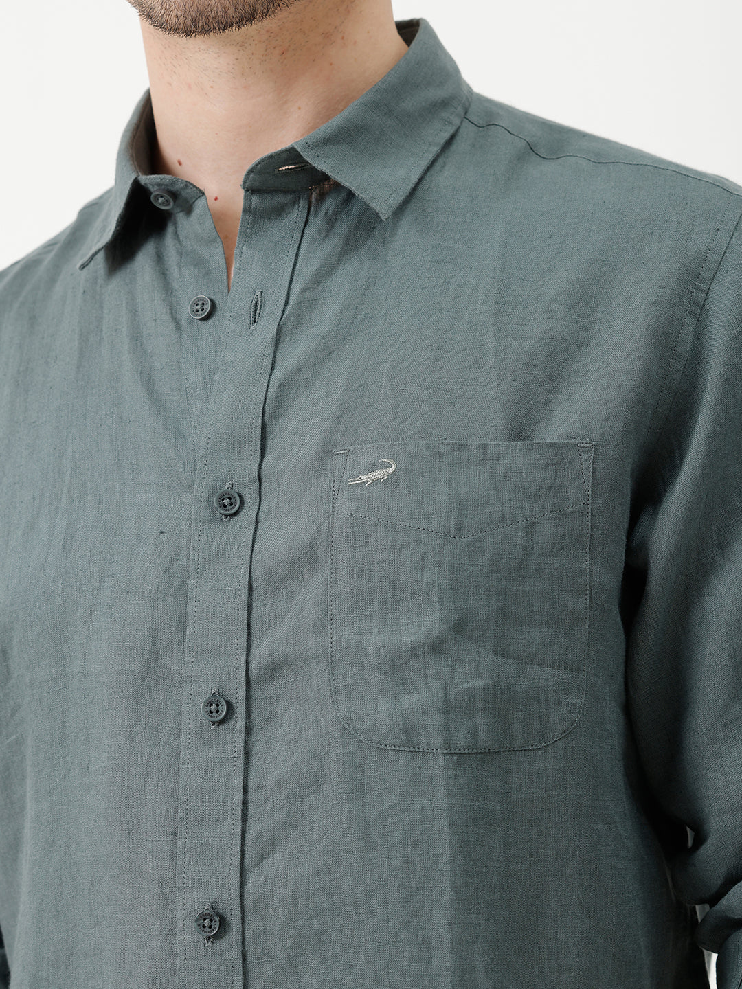 A man wearing a slate blue-green long-sleeve linen button-up shirt with white trousers, standing against a plain white studio background.