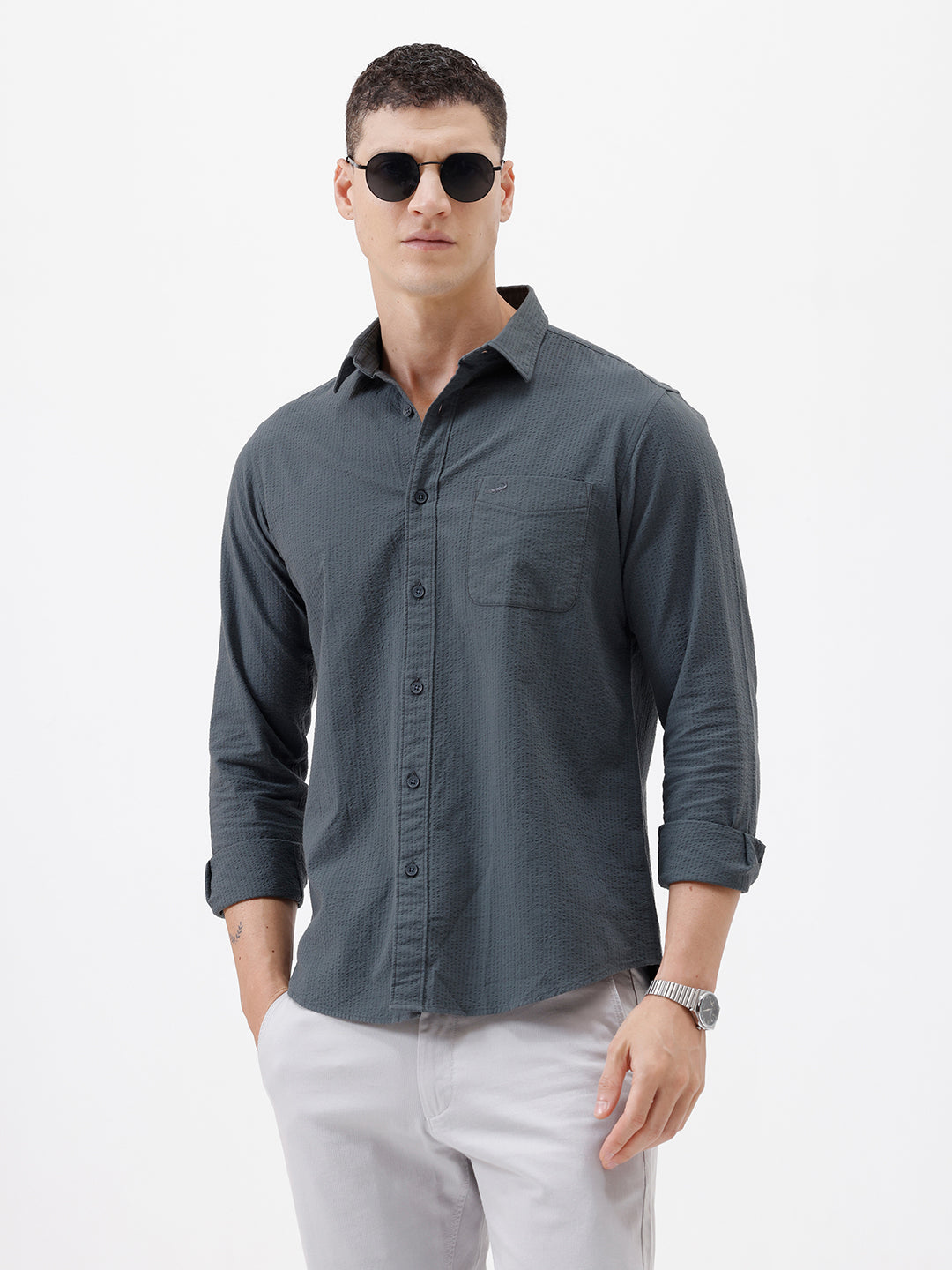 A man wearing a dark charcoal gray long-sleeve button-down shirt with a chest pocket, paired with light gray trousers, standing against a plain white studio background.