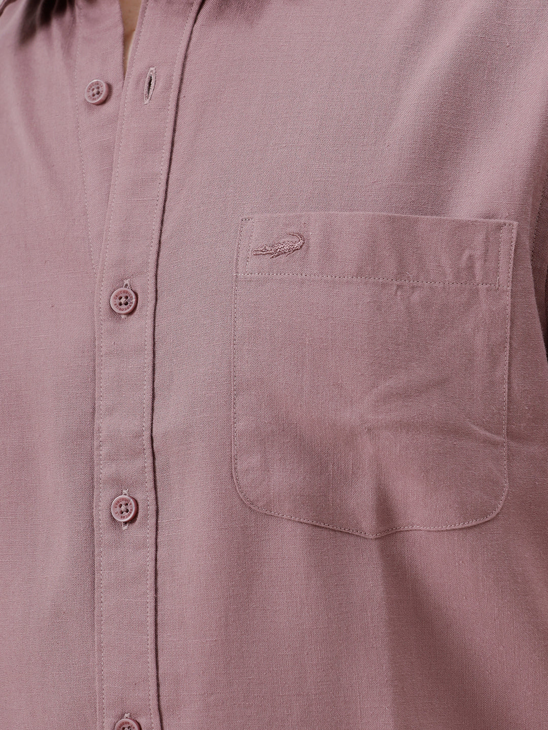 A man wearing a short-sleeve button-up shirt in dusty rose pink with dark blue jeans, standing against a plain white studio background.