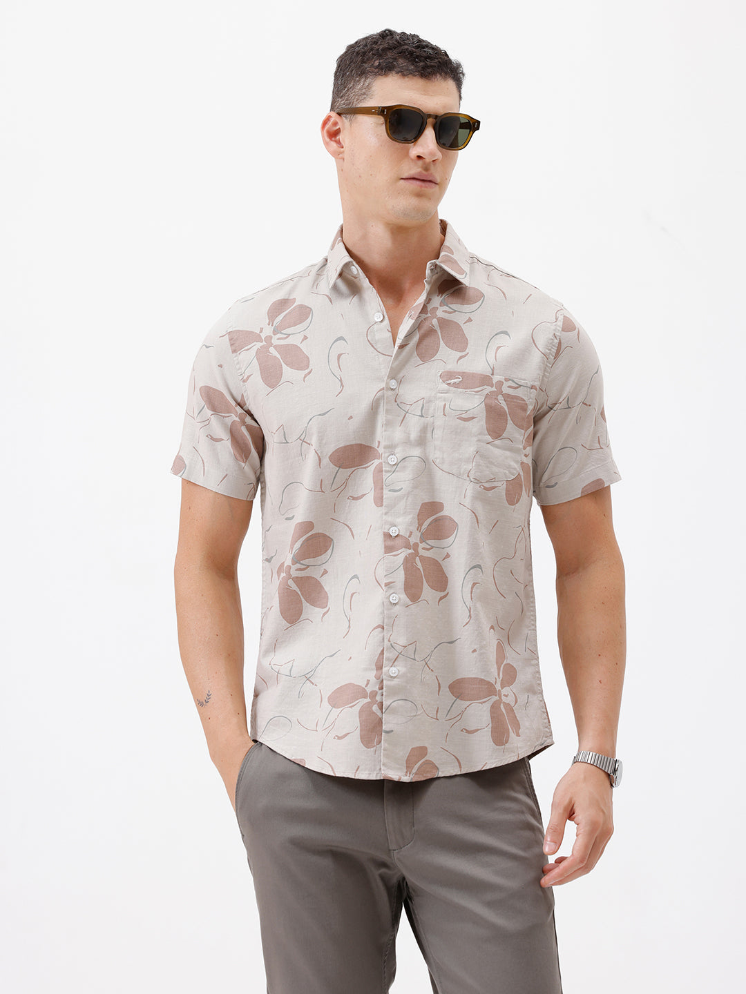 A man wearing a short-sleeve button-up shirt with a beige and brown floral pattern, paired with gray trousers, standing against a plain white background.