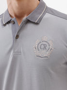 A man wearing a light gray short-sleeve polo shirt with darker gray shoulder panels and a small crest logo on the left chest, paired with khaki cargo pants, against a plain light gray studio background.