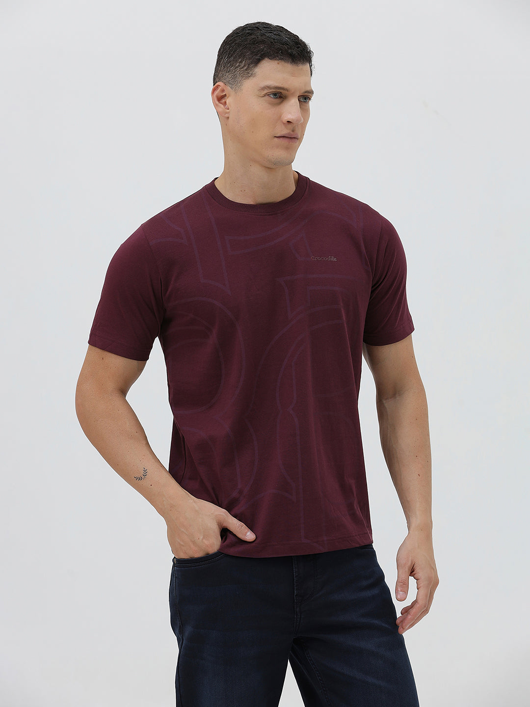 A man wearing a short-sleeve crewneck t-shirt in maroon with a subtle tonal graphic pattern, paired with dark blue jeans, against a light gray studio background.