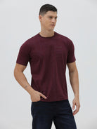 A man wearing a short-sleeve crewneck t-shirt in maroon with a subtle tonal graphic pattern, paired with dark blue jeans, against a light gray studio background.