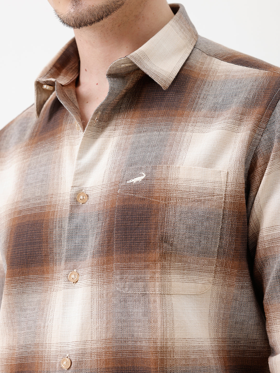 A man wearing a long-sleeve button-up plaid shirt in brown, beige, and gray tones, paired with dark jeans, standing against a plain white studio background.