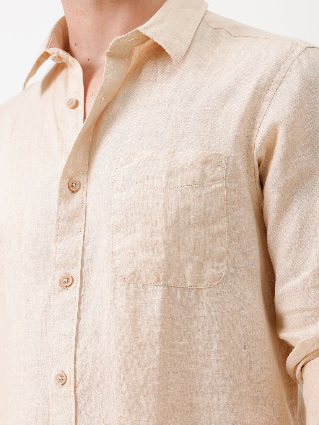 A man wearing a light beige linen button-up shirt with rolled-up sleeves and dark jeans, standing against a plain white studio background.