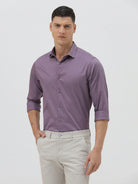 A man wearing a purple long-sleeve button-down shirt with rolled-up sleeves and light beige trousers, standing against a plain light gray background.