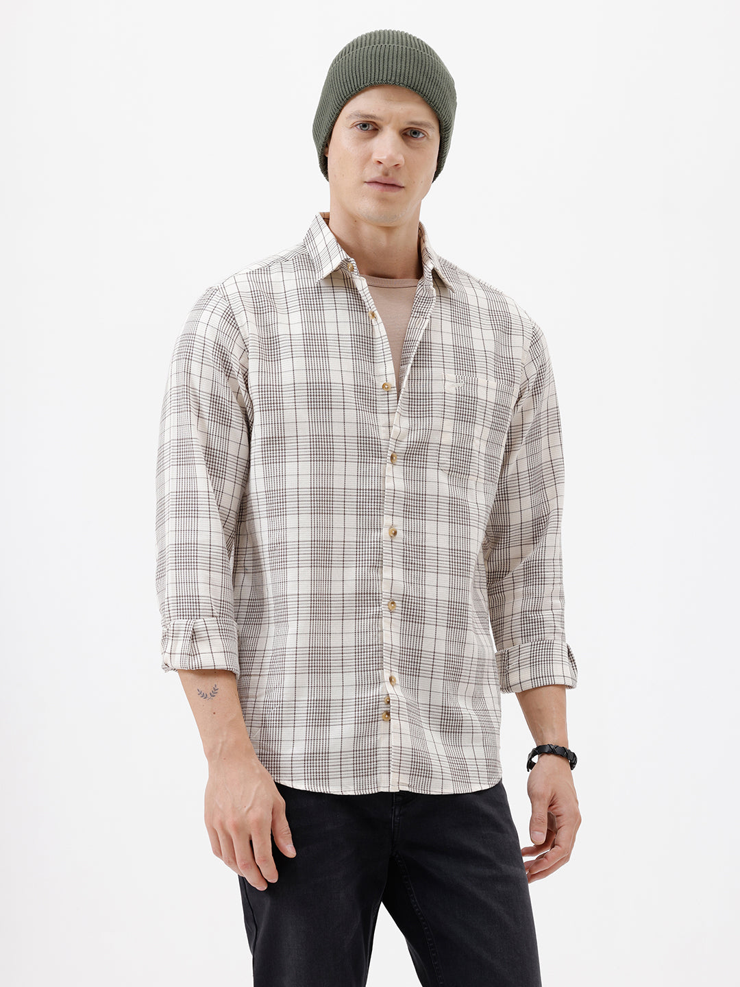 A man wearing a long-sleeve button-up plaid shirt in beige, white, and gray, paired with dark pants, standing against a plain white studio background.