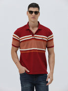 A man wearing a red short-sleeve polo shirt with horizontal cream and rust-orange stripes across the chest and sleeves, paired with dark blue jeans and black sunglasses, against a light gray studio background.