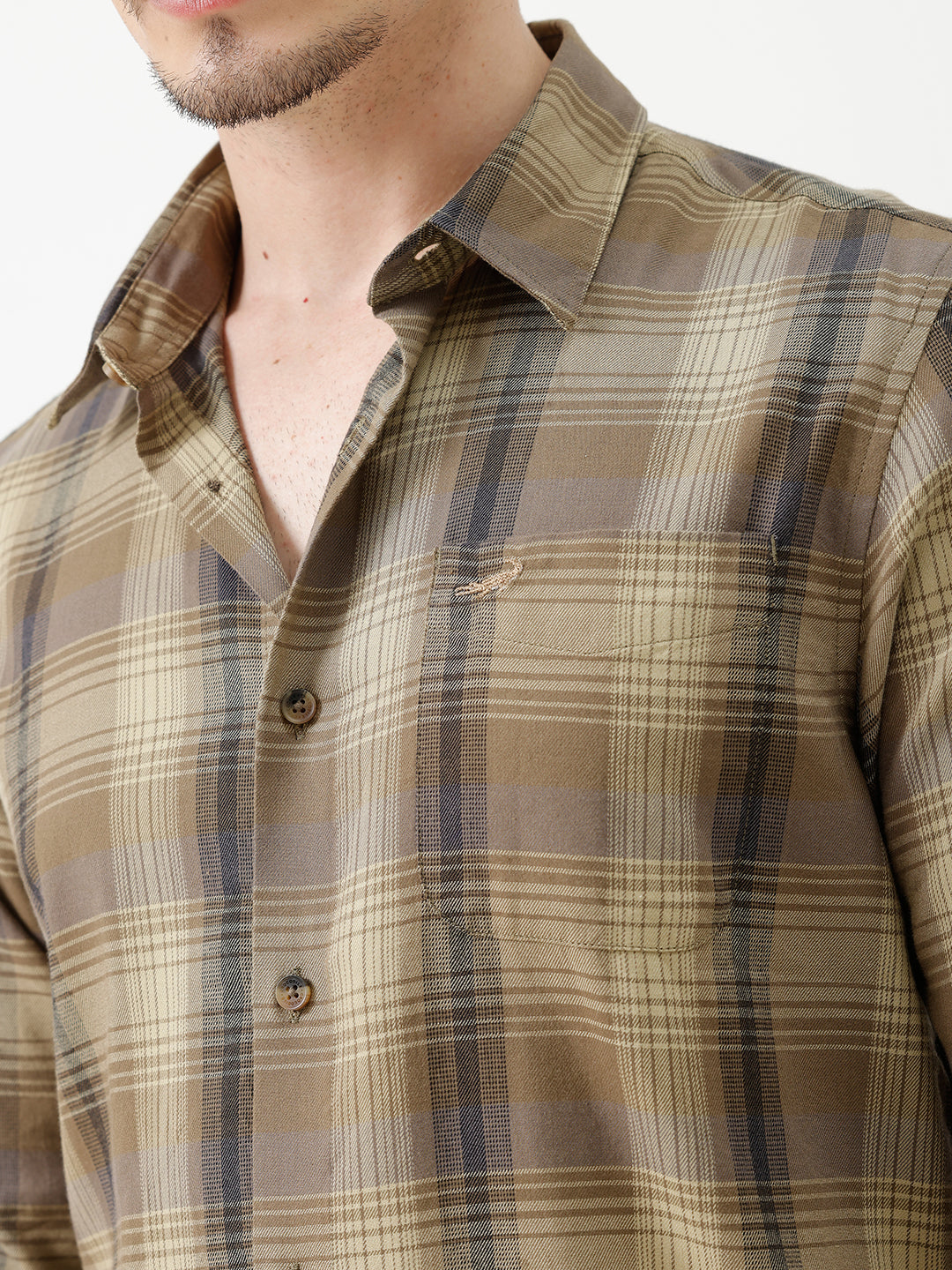 A man wearing a brown and beige plaid long-sleeve button-down shirt with dark sunglasses and black jeans, standing against a plain white studio background.