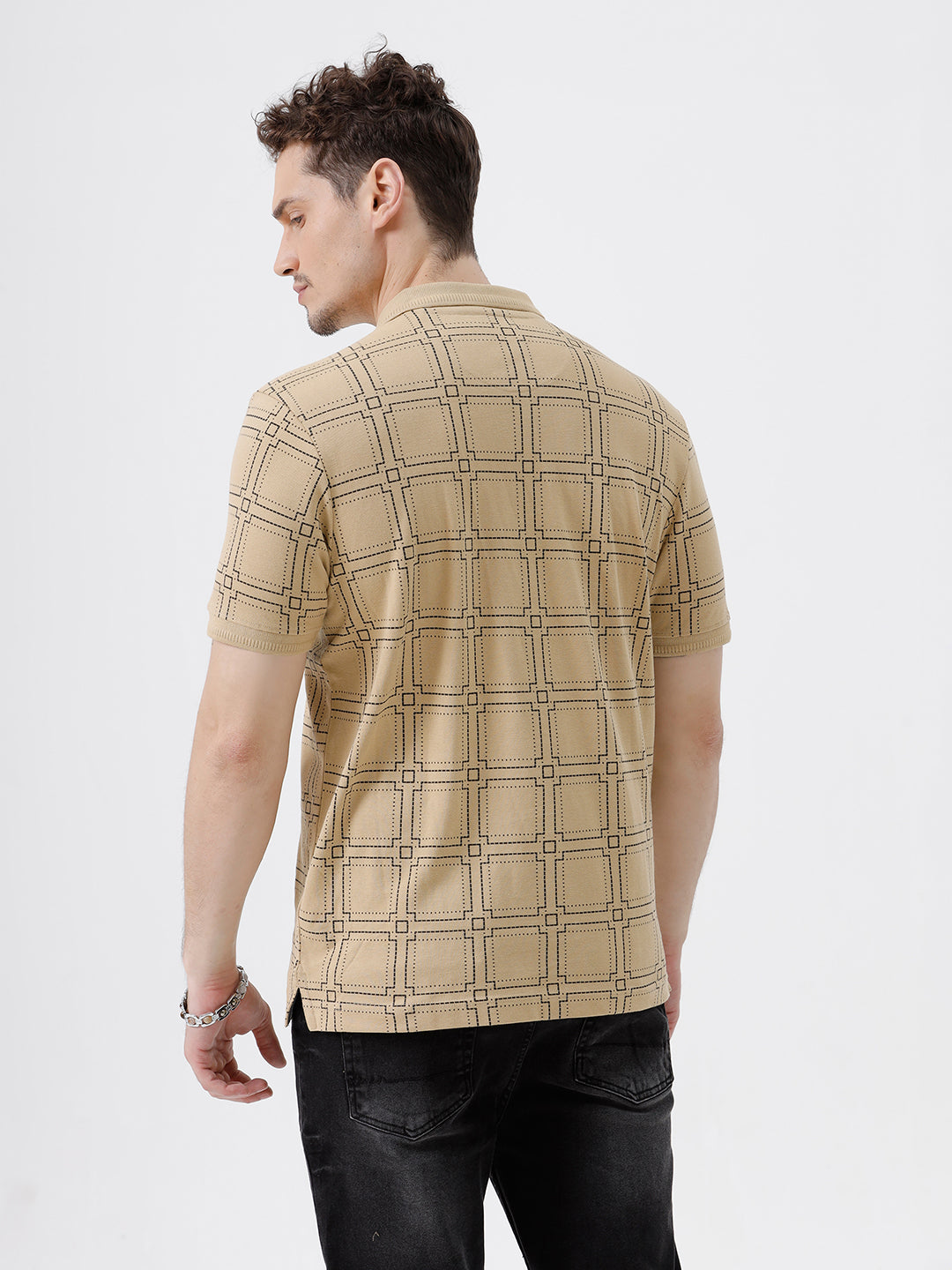 A man wearing a beige short-sleeve polo shirt with a brown grid pattern, paired with black jeans, standing against a plain white studio background.