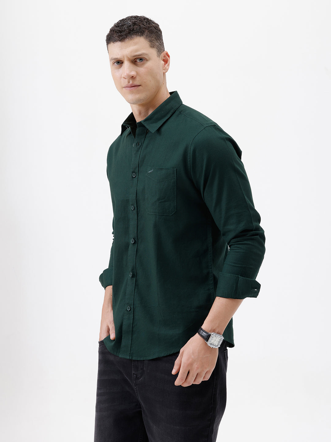 A man wearing a dark forest green long-sleeve button-down shirt with a chest pocket, paired with dark jeans, standing against a plain white studio background.