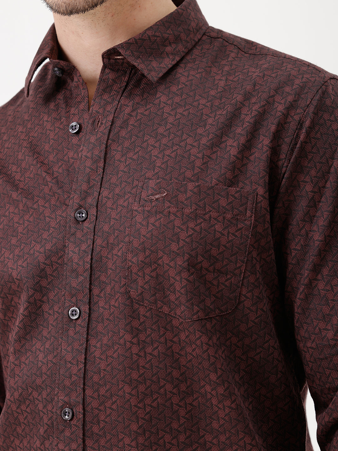 A man wearing a dark burgundy long-sleeve button-up shirt with a subtle geometric pattern, paired with beige trousers, standing against a plain white studio background.