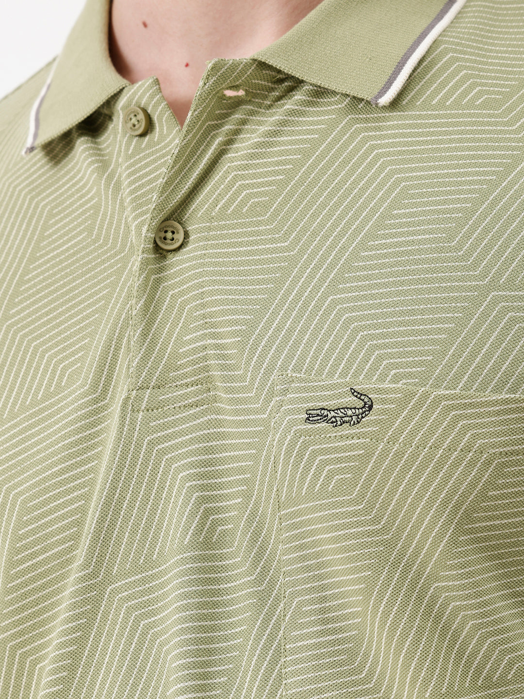 A man wearing a light sage green short-sleeve polo shirt with a subtle geometric pattern, paired with beige trousers, standing against a plain white studio background.