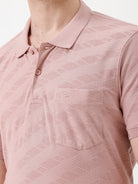A man wearing a dusty pink short-sleeve polo shirt with a chest pocket, paired with light beige trousers, standing against a plain white studio background.