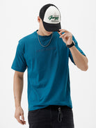 A man wearing a teal blue short-sleeve t-shirt with a subtle tonal 'GREATNESS' text graphic across the chest, paired with black pants, photographed against a clean white studio background.