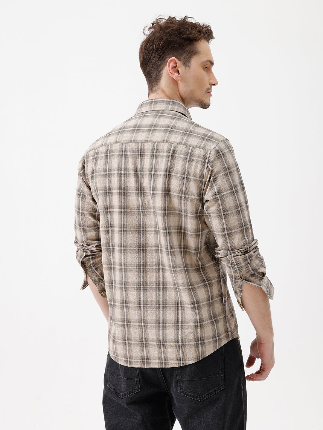 A man wearing a long-sleeve button-up plaid shirt in beige and brown tones, paired with dark jeans, standing against a plain white studio background.