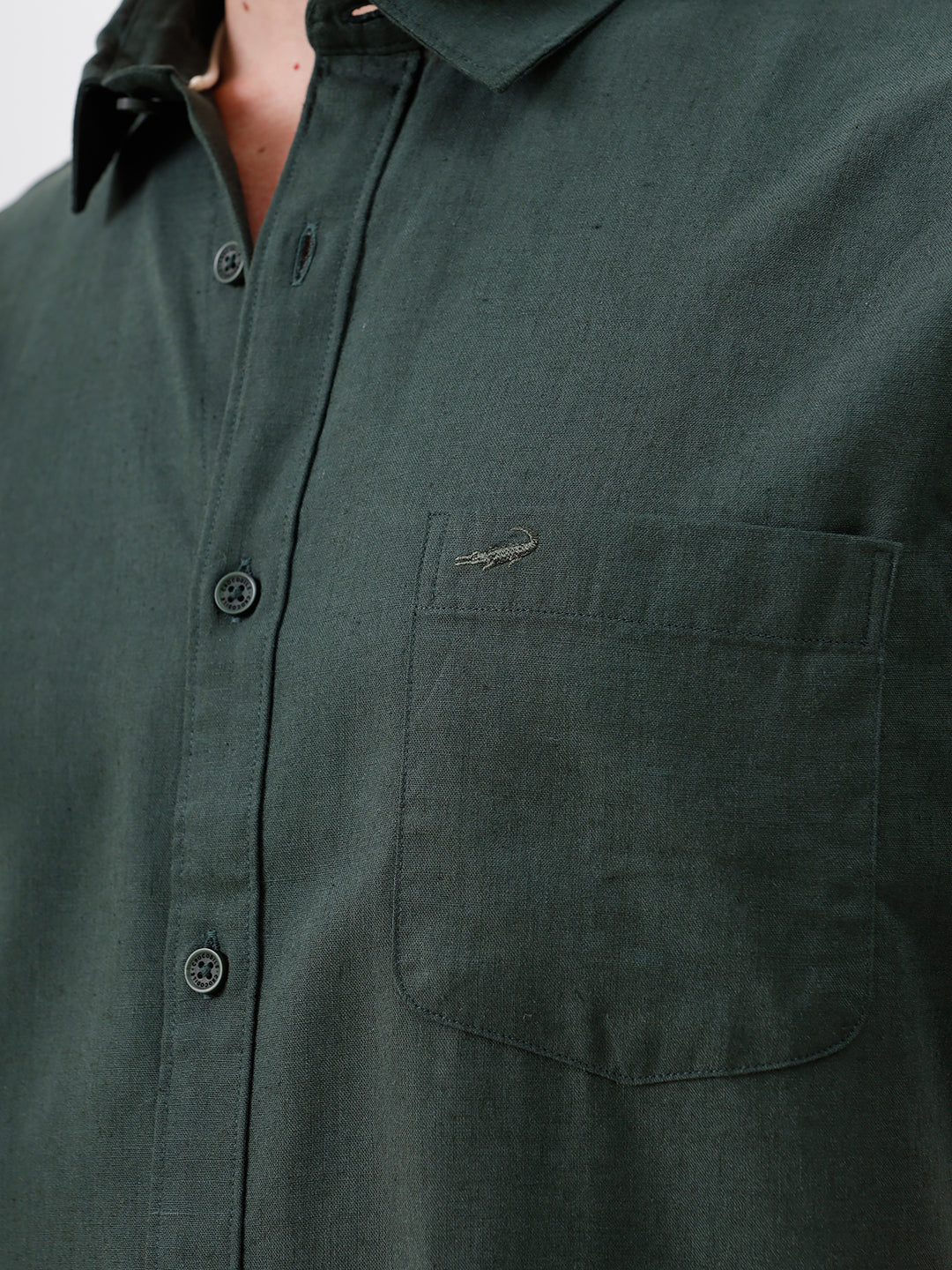 A man wearing a dark forest green short-sleeve button-up shirt with a chest pocket, paired with black jeans, standing against a plain white studio background.