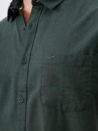 A man wearing a dark forest green short-sleeve button-up shirt with a chest pocket, paired with black jeans, standing against a plain white studio background.