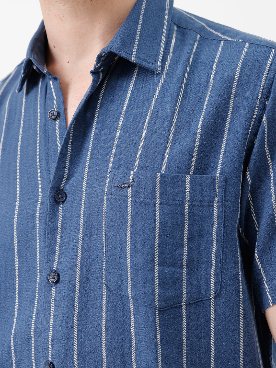 A man wearing a blue short-sleeve button-up shirt with thin white vertical stripes and beige trousers, standing against a plain white studio background.