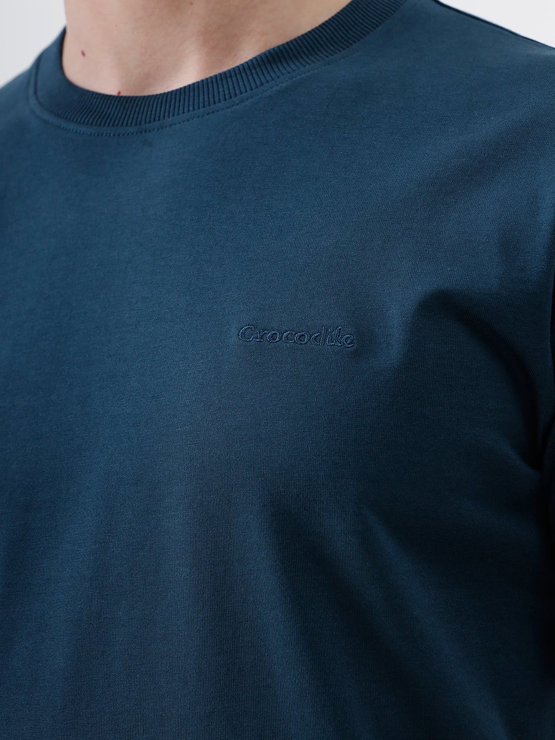 A man wearing a dark navy blue crew-neck t-shirt and black jeans, standing against a light gray studio background.