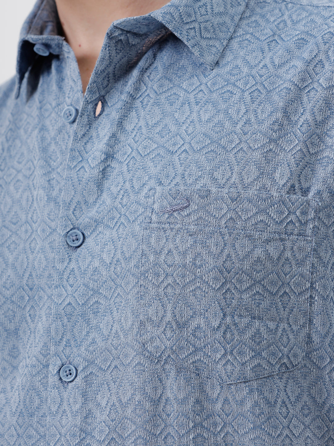 A man wearing a short-sleeve button-up shirt with a fine blue and white geometric pattern, paired with light beige trousers, standing against a plain light gray background.