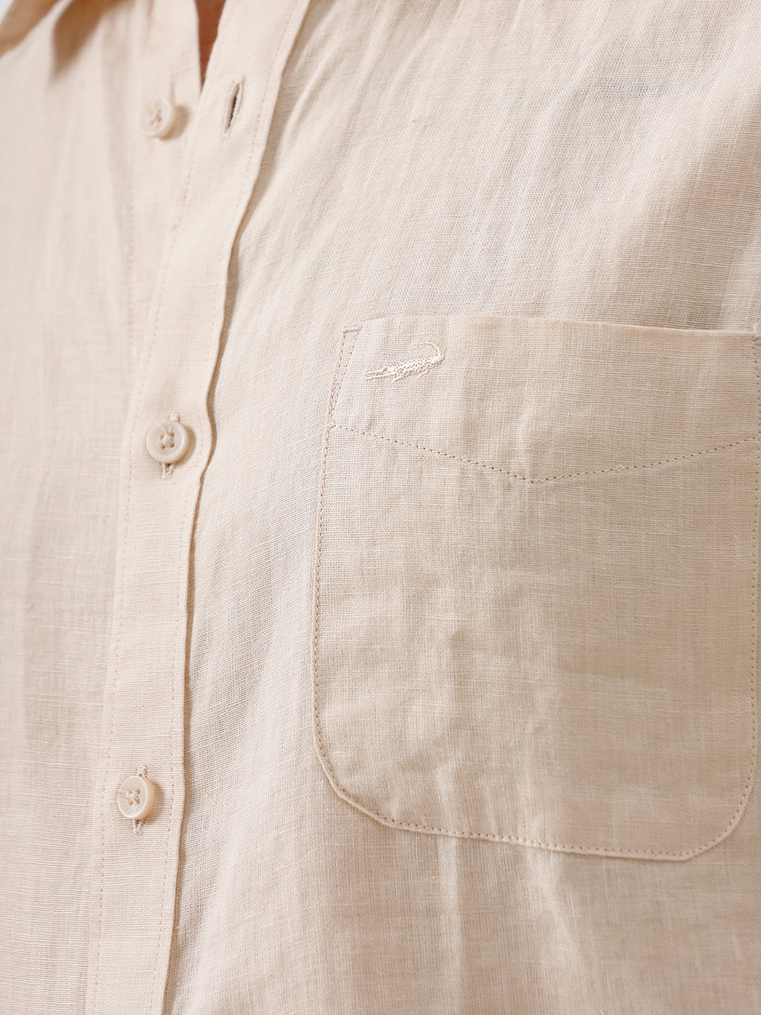 A man wearing a light beige short-sleeve linen button-up shirt and taupe trousers, standing against a plain white studio background.