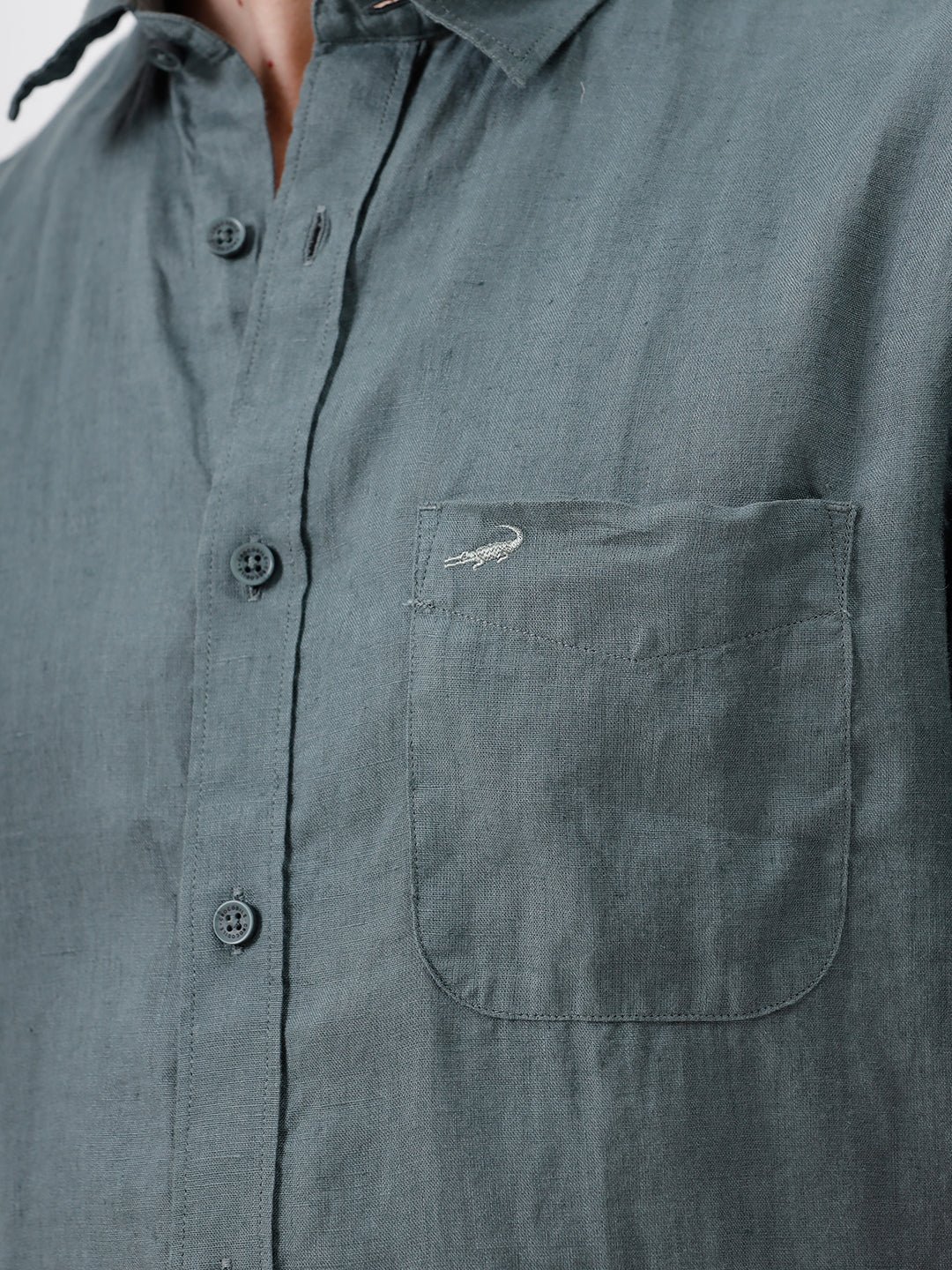 A man wearing a short-sleeve button-up shirt in a muted slate green color, paired with light beige trousers, standing against a plain light gray background.