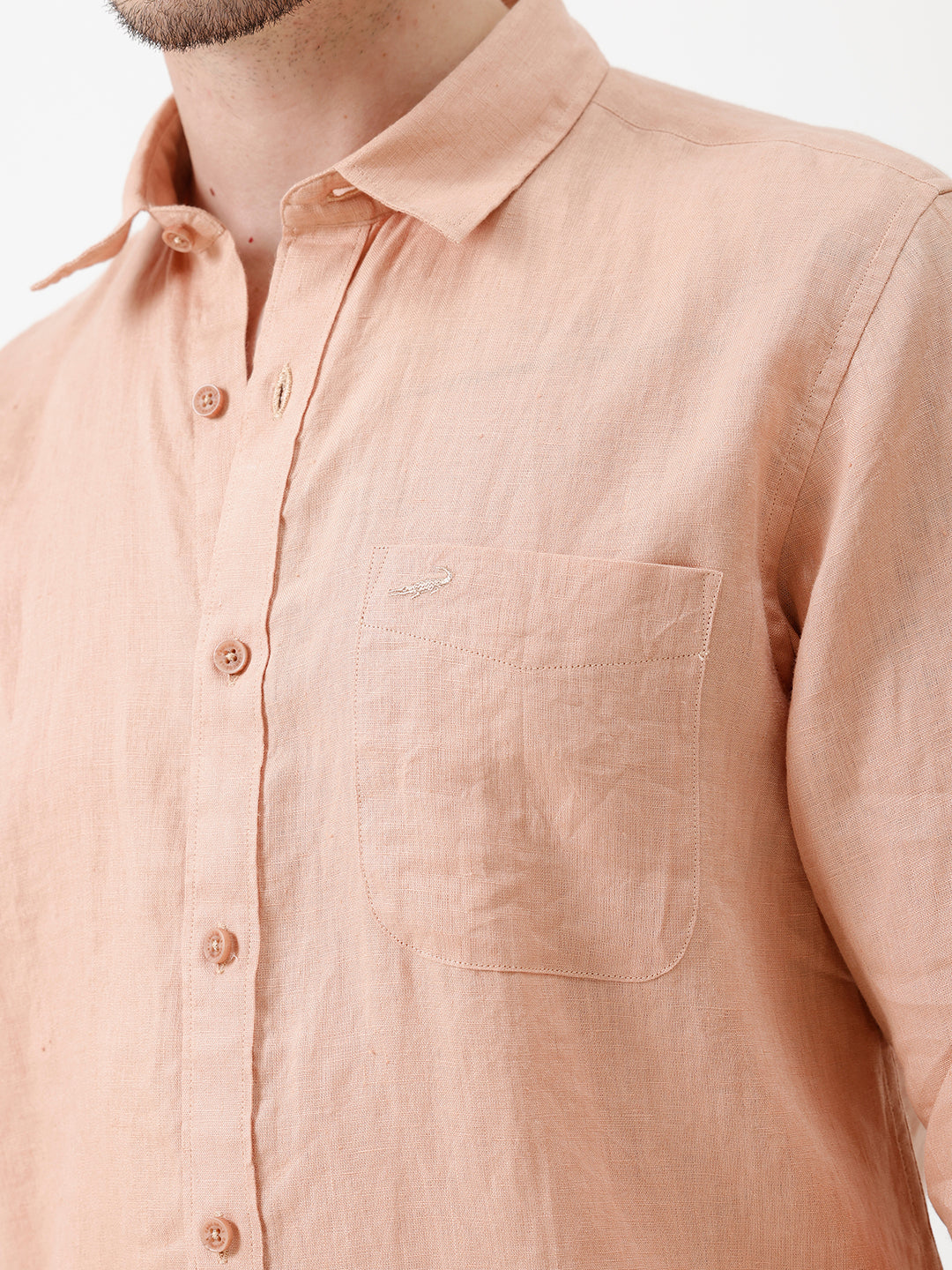 A man wearing a light peach-colored linen button-down shirt with dark jeans, standing against a plain white studio background.