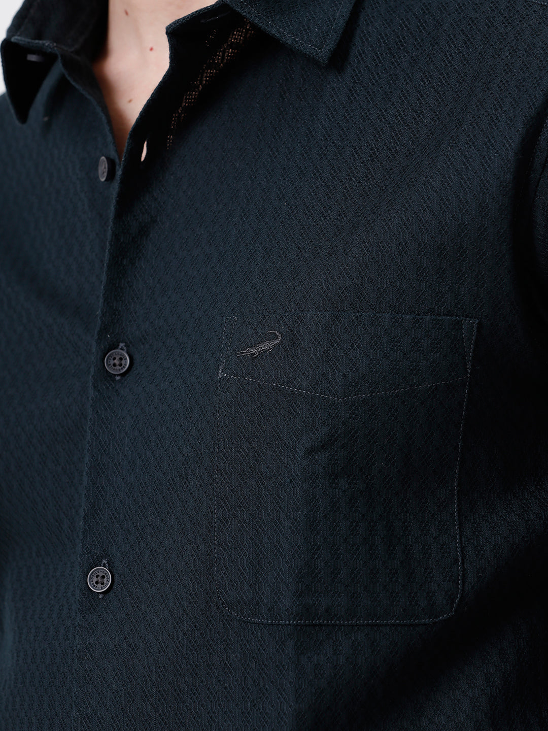A man wearing a dark navy long-sleeve button-up shirt with a subtle textured pattern and light beige trousers, standing against a plain light gray background.