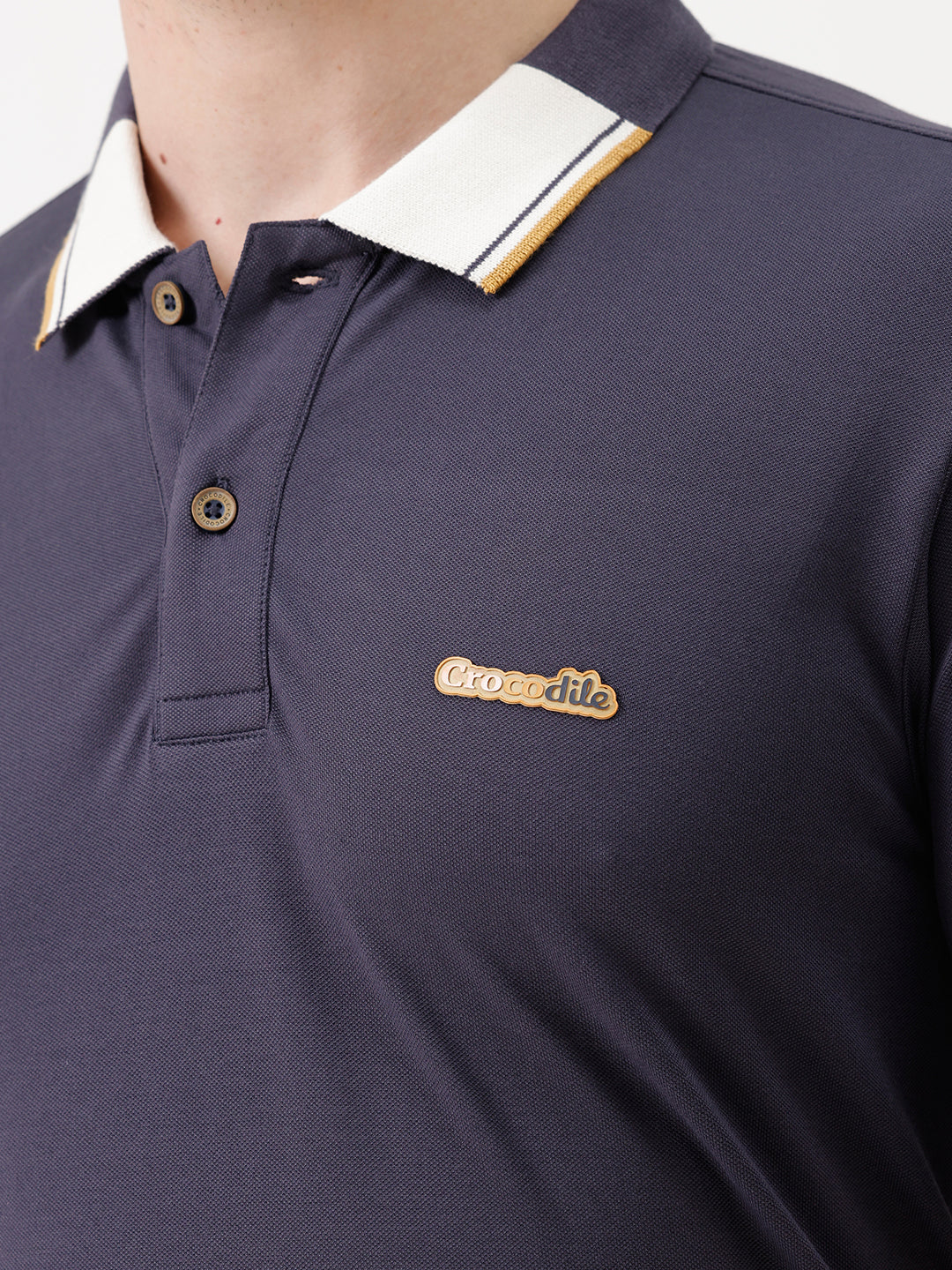 A man wearing a navy blue short-sleeve polo shirt with a contrasting cream and gold striped collar, paired with beige trousers, against a plain white studio background.