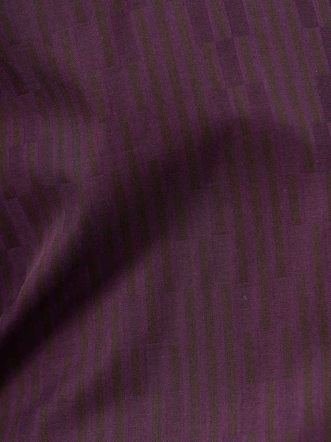 A man wearing a dark purple long-sleeve button-down shirt with subtle vertical stripes and dark navy trousers, standing against a light gray background.