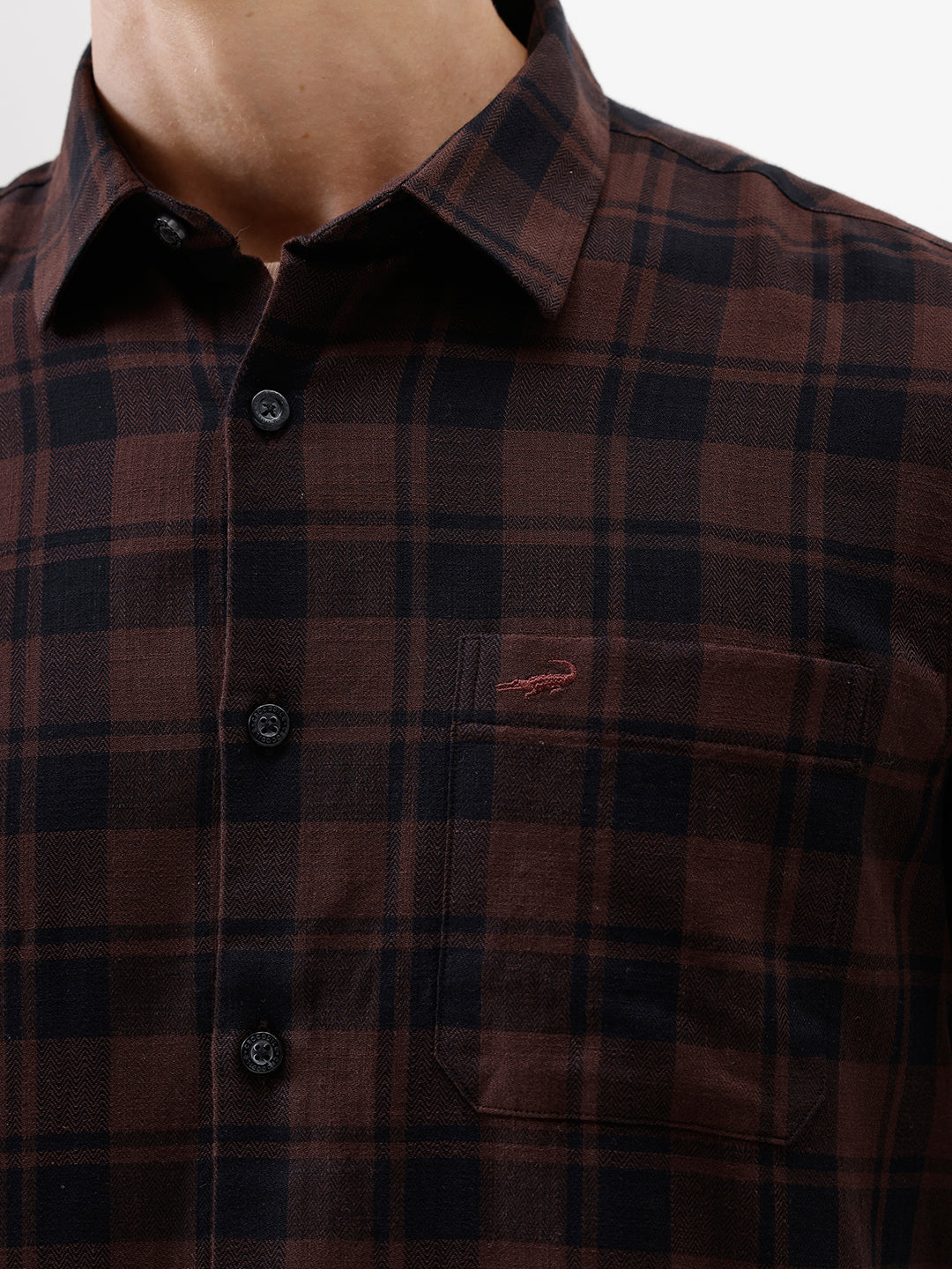 A man wearing a dark brown and black plaid long-sleeve button-up shirt, paired with dark pants, standing against a plain light gray background.