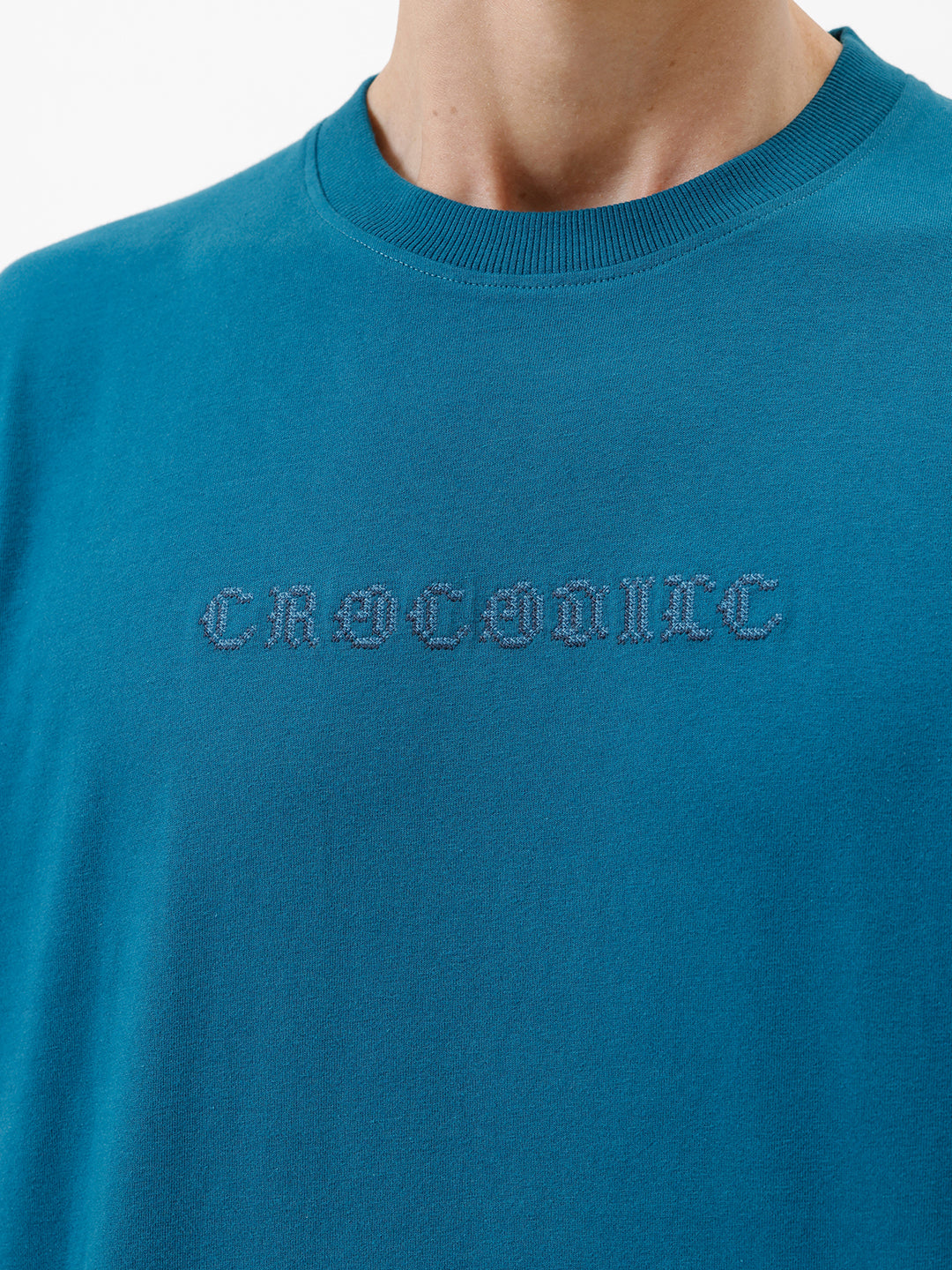 A man wearing a teal blue short-sleeve t-shirt with a subtle tonal 'GREATNESS' text graphic across the chest, paired with black pants, photographed against a clean white studio background.