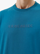 A man wearing a teal blue short-sleeve t-shirt with a subtle tonal 'GREATNESS' text graphic across the chest, paired with black pants, photographed against a clean white studio background.