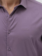 A man wearing a purple long-sleeve button-down shirt with rolled-up sleeves and light beige trousers, standing against a plain light gray background.