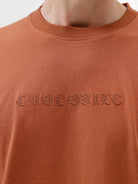 A man wearing a rust-orange crewneck t-shirt with a subtle tonal 'ELECTRIC' logo across the chest, paired with black pants, photographed against a clean white studio background.
