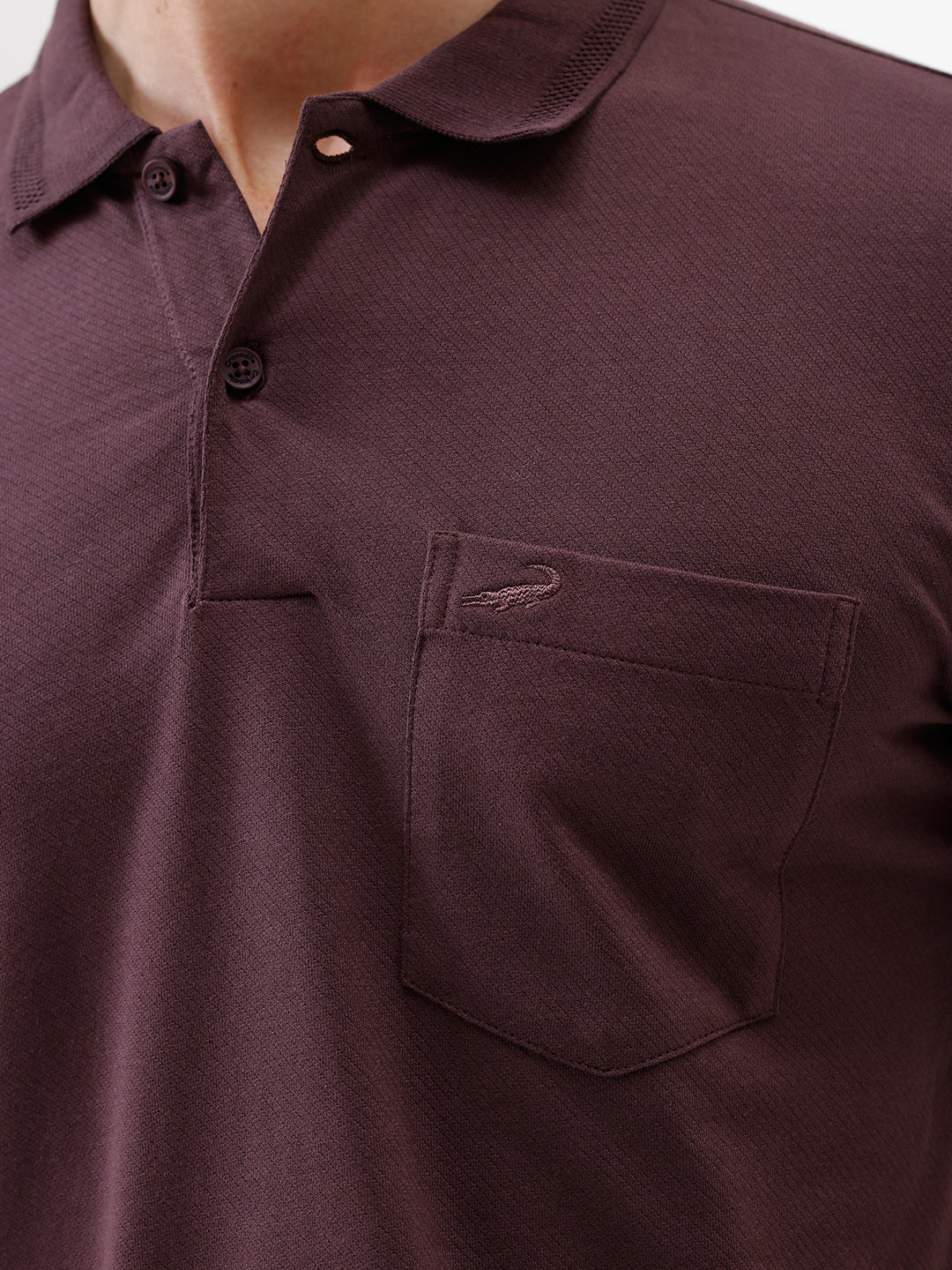 A man wearing a short-sleeve polo shirt in dark burgundy with a chest pocket, paired with gray trousers, standing against a plain light gray background.