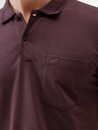 A man wearing a short-sleeve polo shirt in dark burgundy with a chest pocket, paired with gray trousers, standing against a plain light gray background.